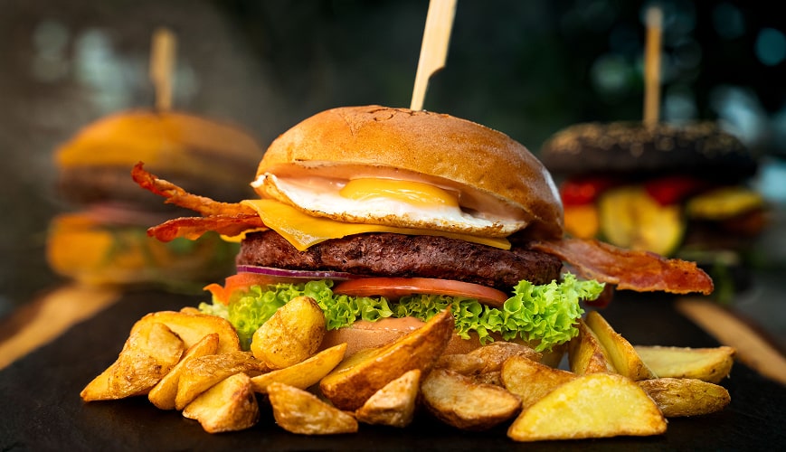 Hamburger z chrupiącym bekonem, świeżą sałatą i dojrzałym pomidorem, podany z grillowanymi ziemniakami na eleganckim czarnym talerzu.