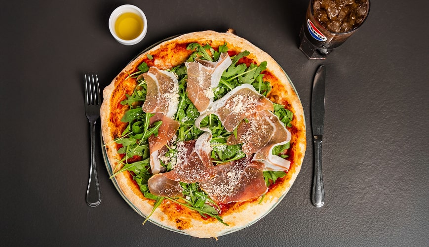Włoska pizza z dojrzewającą szynką, rukolą i parmezanem podana z oliwą i colą na czarnej tacy.
